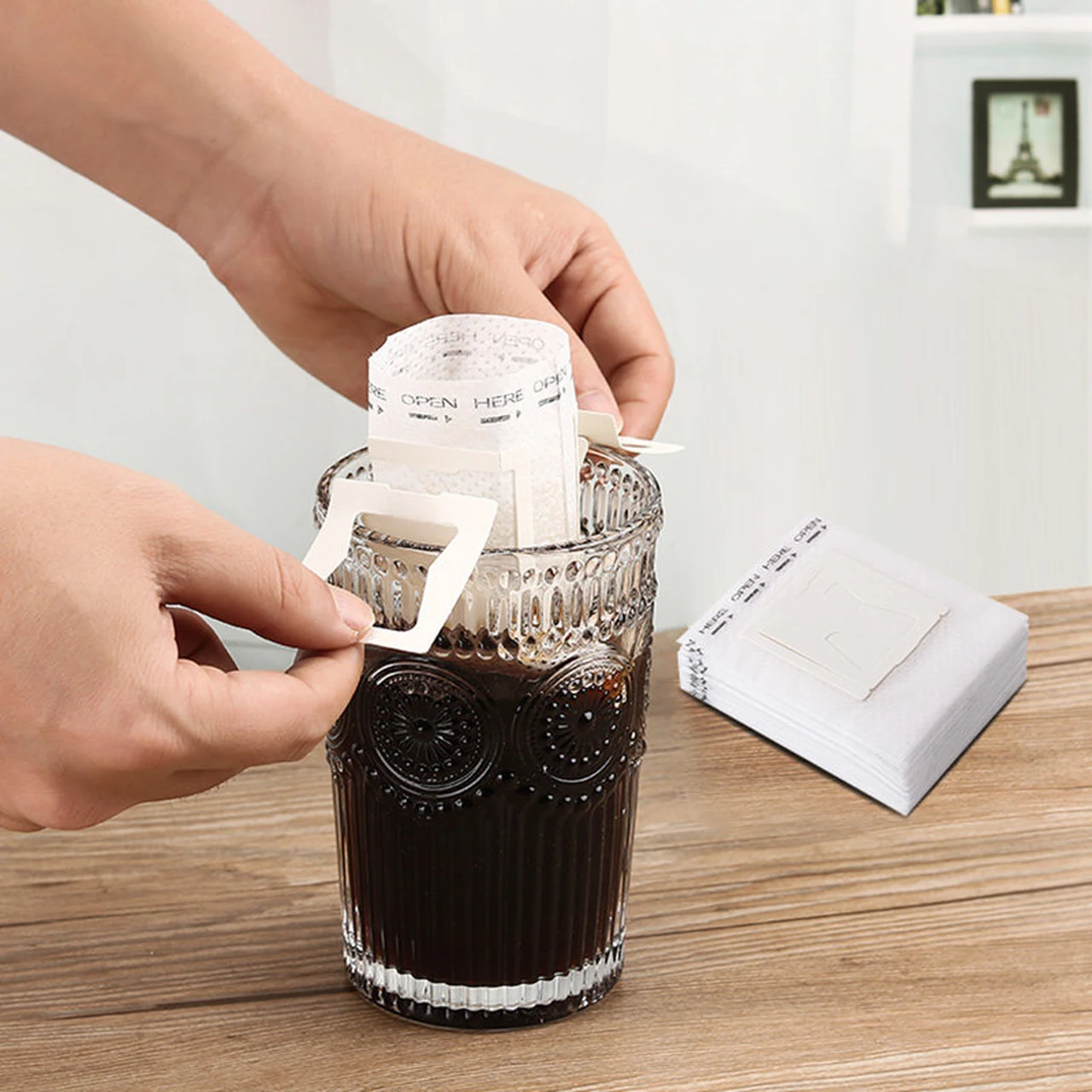 Hands opening a drip coffee filter bag and placing its supports over a glass.