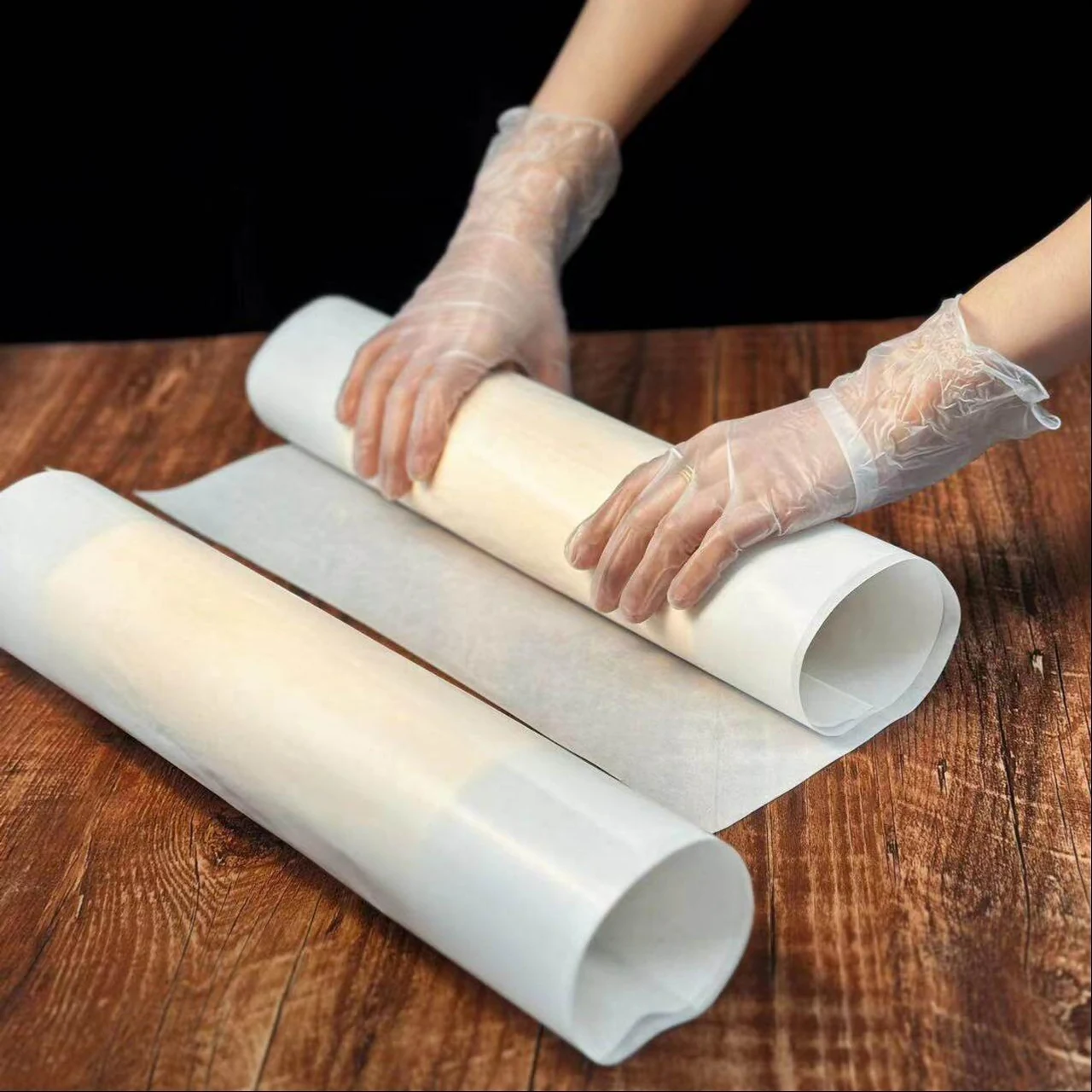 Gloved hands unrolling white baking paper on a wooden worktop.